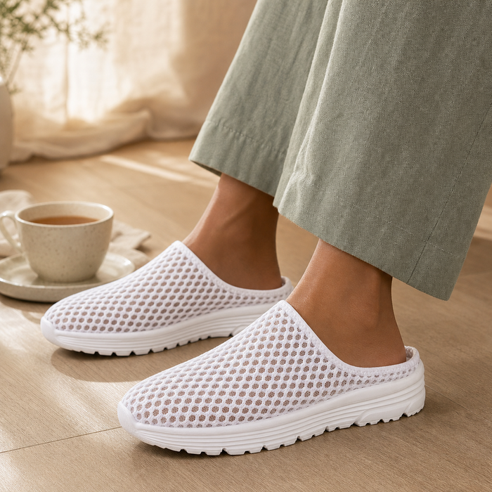 White Airomi mesh mules worn indoors on a wood floor beside a cup of tea