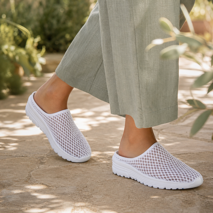 White Airomi mesh mules worn mid-step on a sunlit garden path